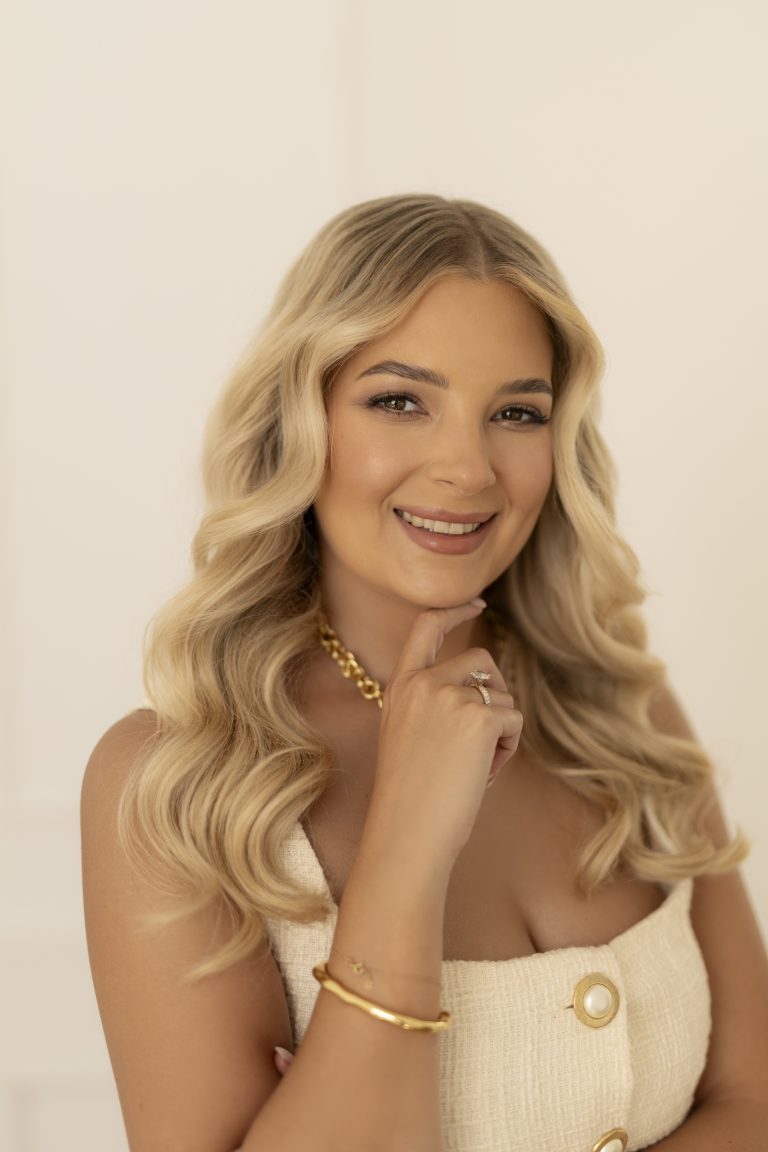 Portret uśmiechniętej kobiety o długich, falowanych blond włosach. Kobieta ma na sobie kremowy top i złotą biżuterię, a dłoń trzyma delikatnie przy brodzie. Tło jest jasne i jednolite.