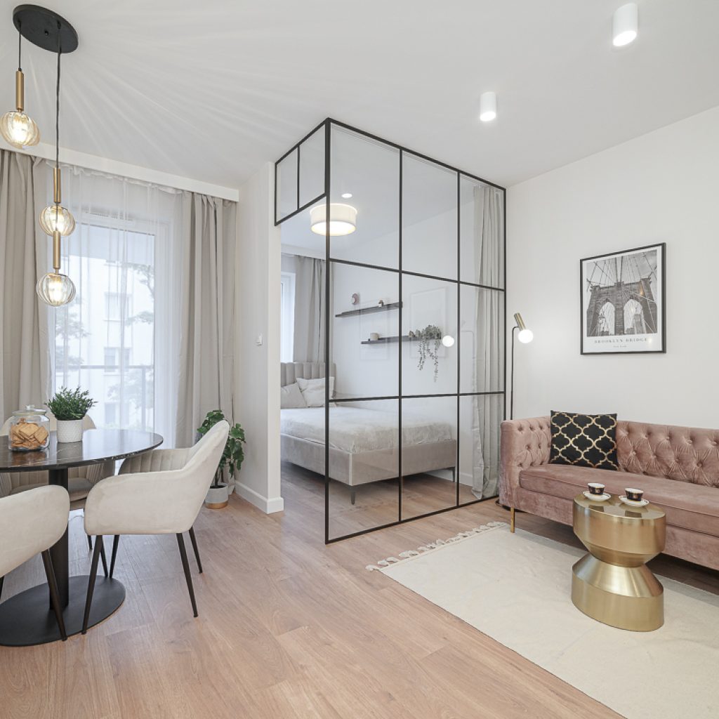 Elegancki salon z welurową, brudnoróżową sofą typu Chesterfield i złotym stolikiem kawowym. Część sypialniana oddzielona jest przeszkloną ścianą z czarnymi szprosami. Po lewej stronie stoi mały, okrągły stół jadalniany z jasnymi fotelami.